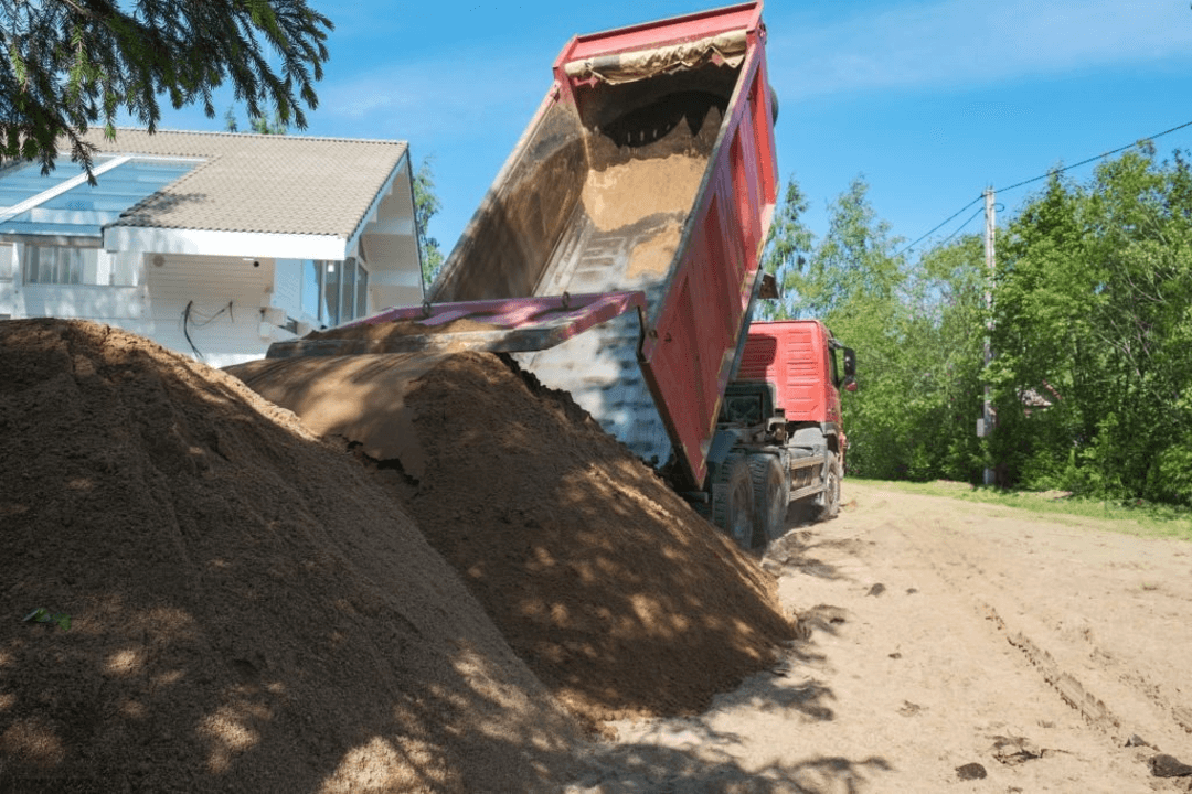 Red dump truck unloading sand next to a residential house with trees and clear sky in the background.