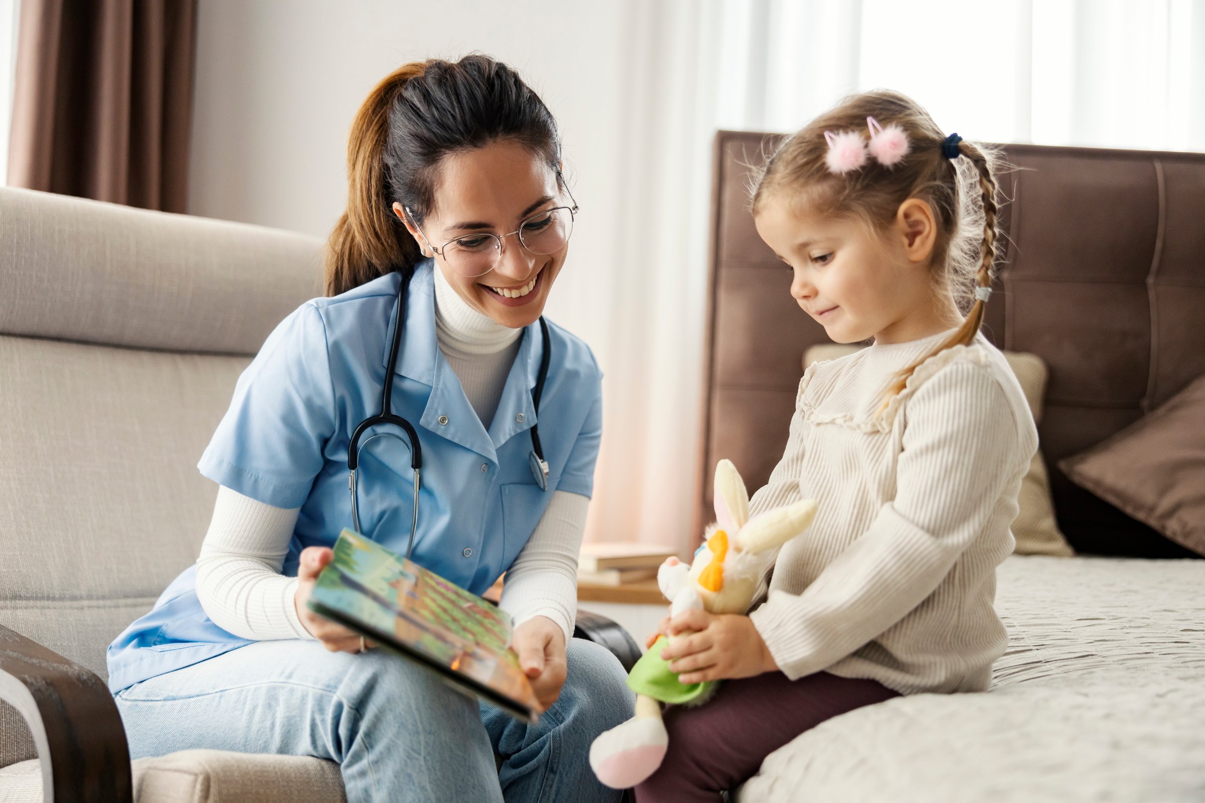 Caring female pediatrician sitting in bedroom with cute little girl and reading a story.