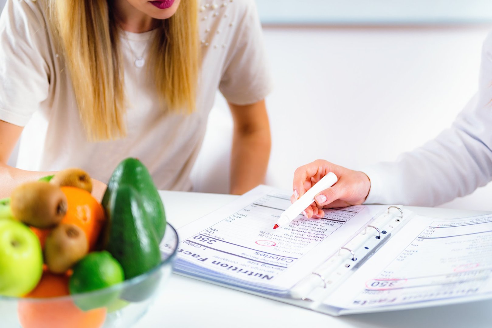 A nutritionist explaining a meal plan to a woman at a table with fruits and vegetables in a bowl nearby.