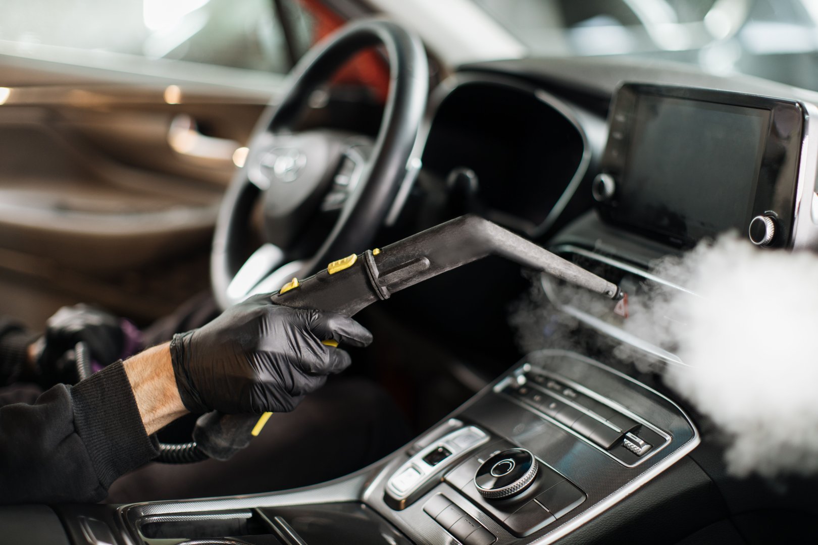 Close up of hands of man in black protective rubber gloves cleaning interior of the car with hot steam cleaner. Selective focus on guy hands. Auto cleaning service and detailing concept.