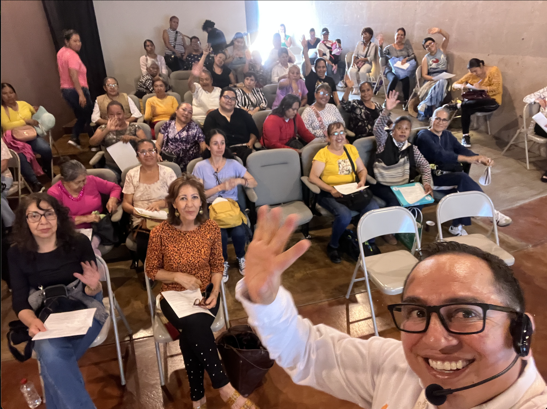 A group of people in a room smiling and waving at the camera, with a person in front taking a selfie.