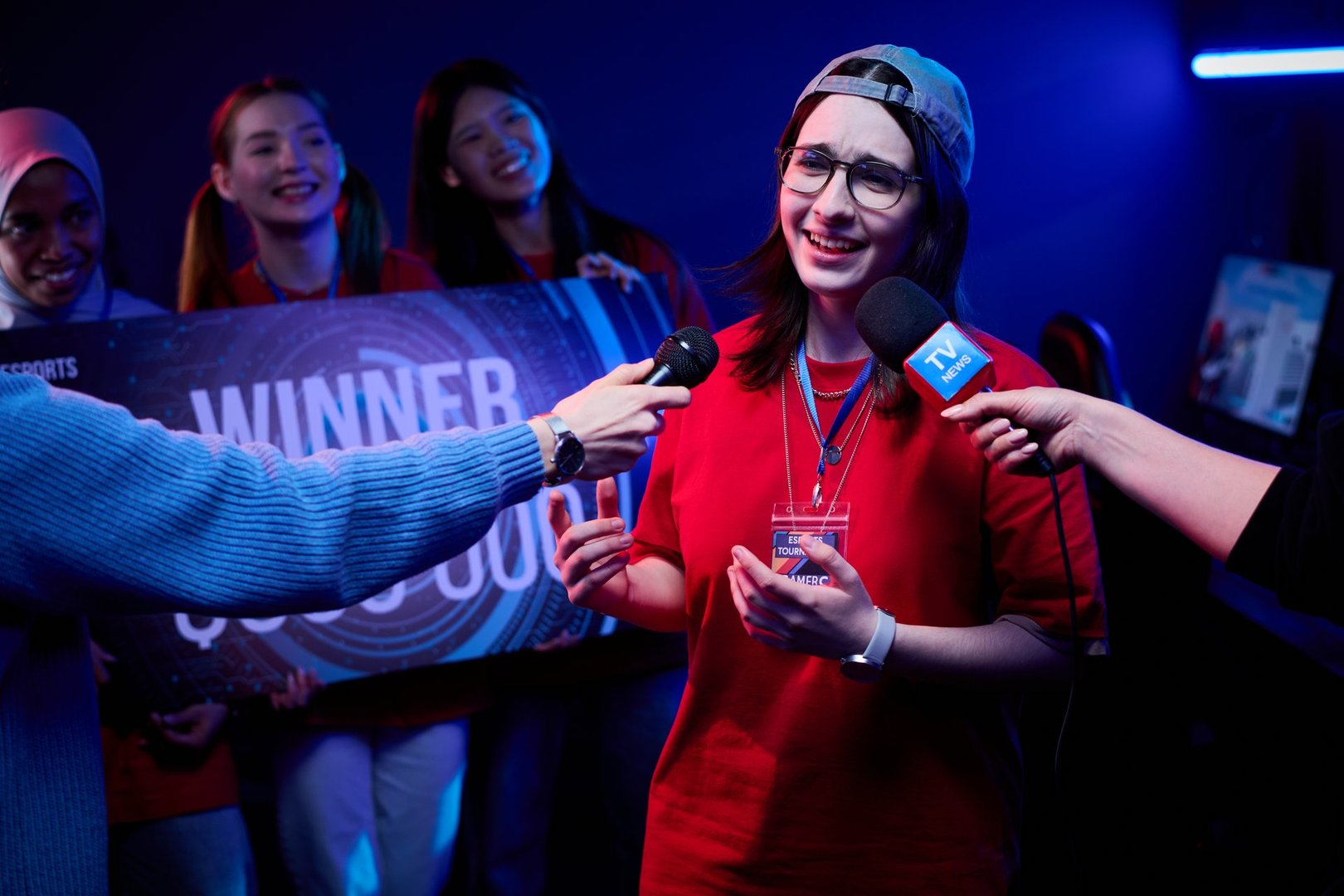 Diverse group of teenage girls celebrating esports victory, Caucasian teenage girl wearing glasses and cap giving interview to reporters while holding trophy, teammates smiling in background