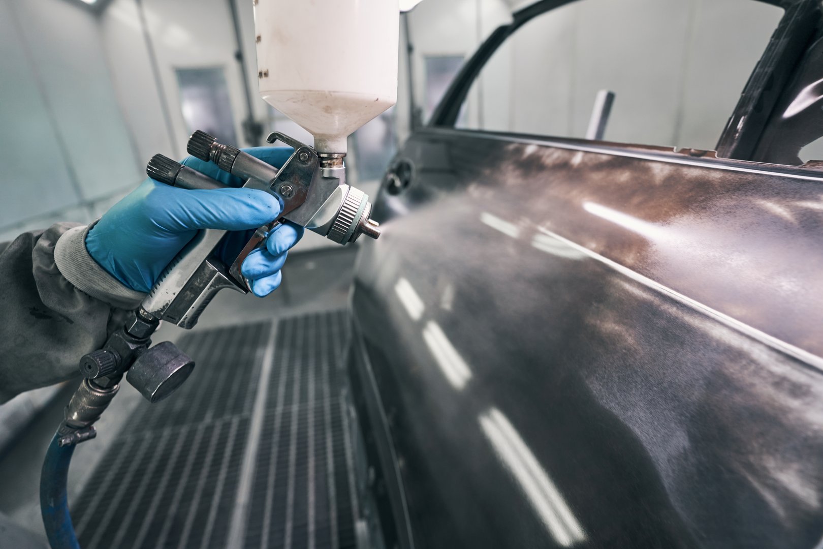 Mechanic dressed in protective overalls and rubber gloves releasing painting spray from gun to car
