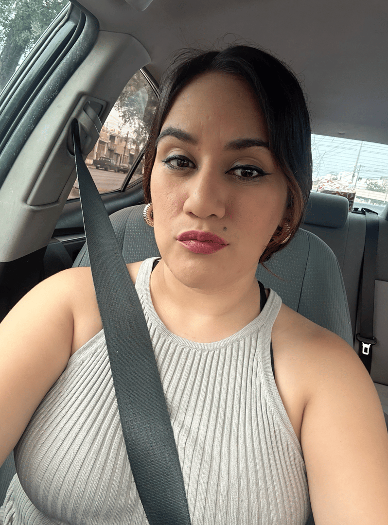 A woman in a sleeveless top sits in a car, wearing a seatbelt. She has dark hair and is looking at the camera.
