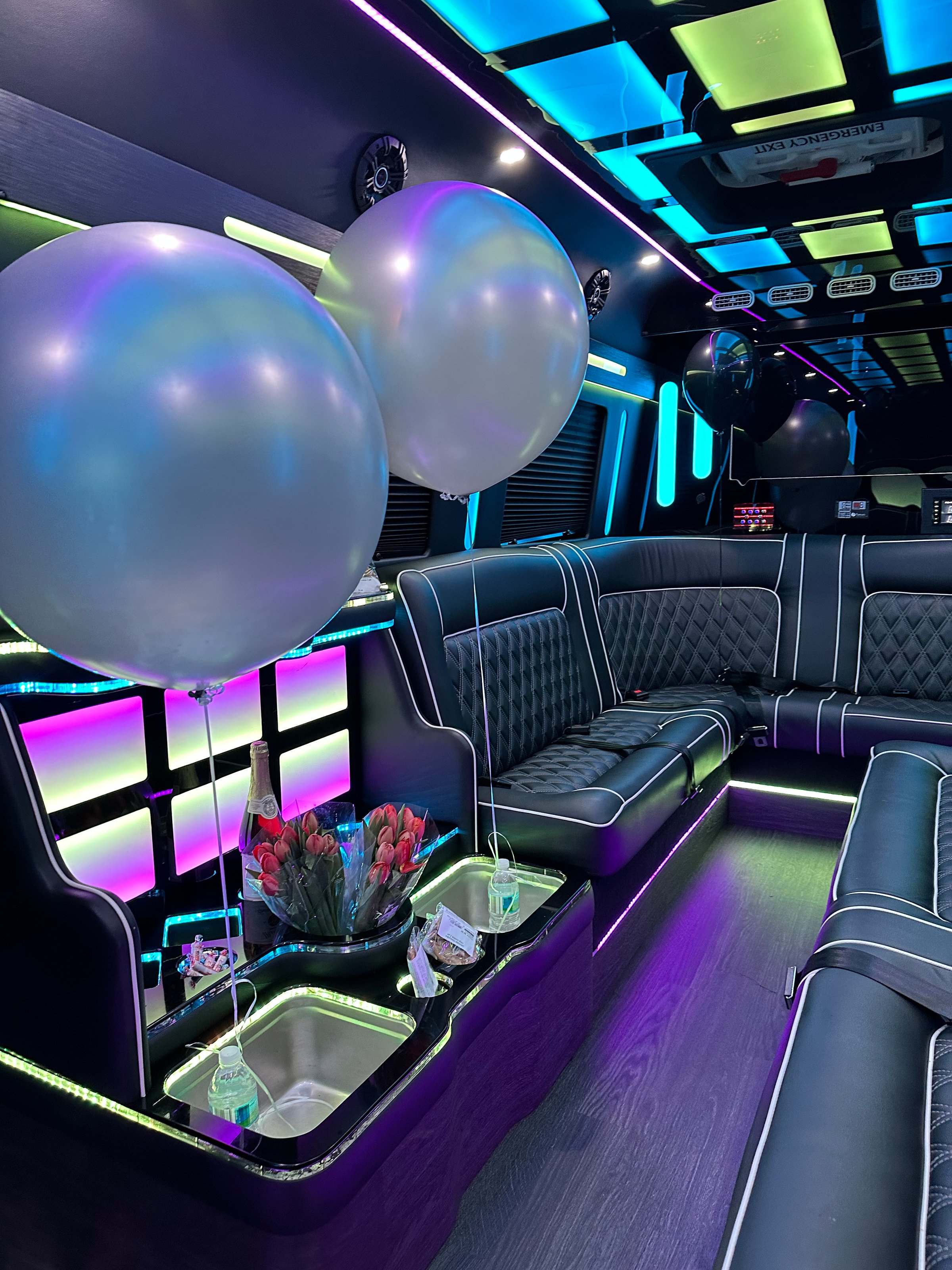 Luxurious party bus interior with neon lights, plush seating, two large balloons, and a bouquet of flowers on a table.