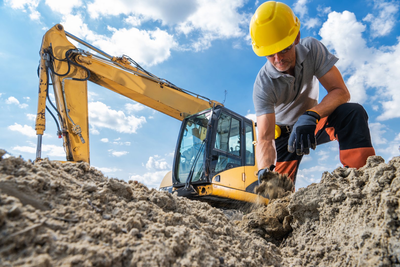 A construction worker is kneeling in the sand, using his hands to dig while an excavator operates nearby under clear skies.