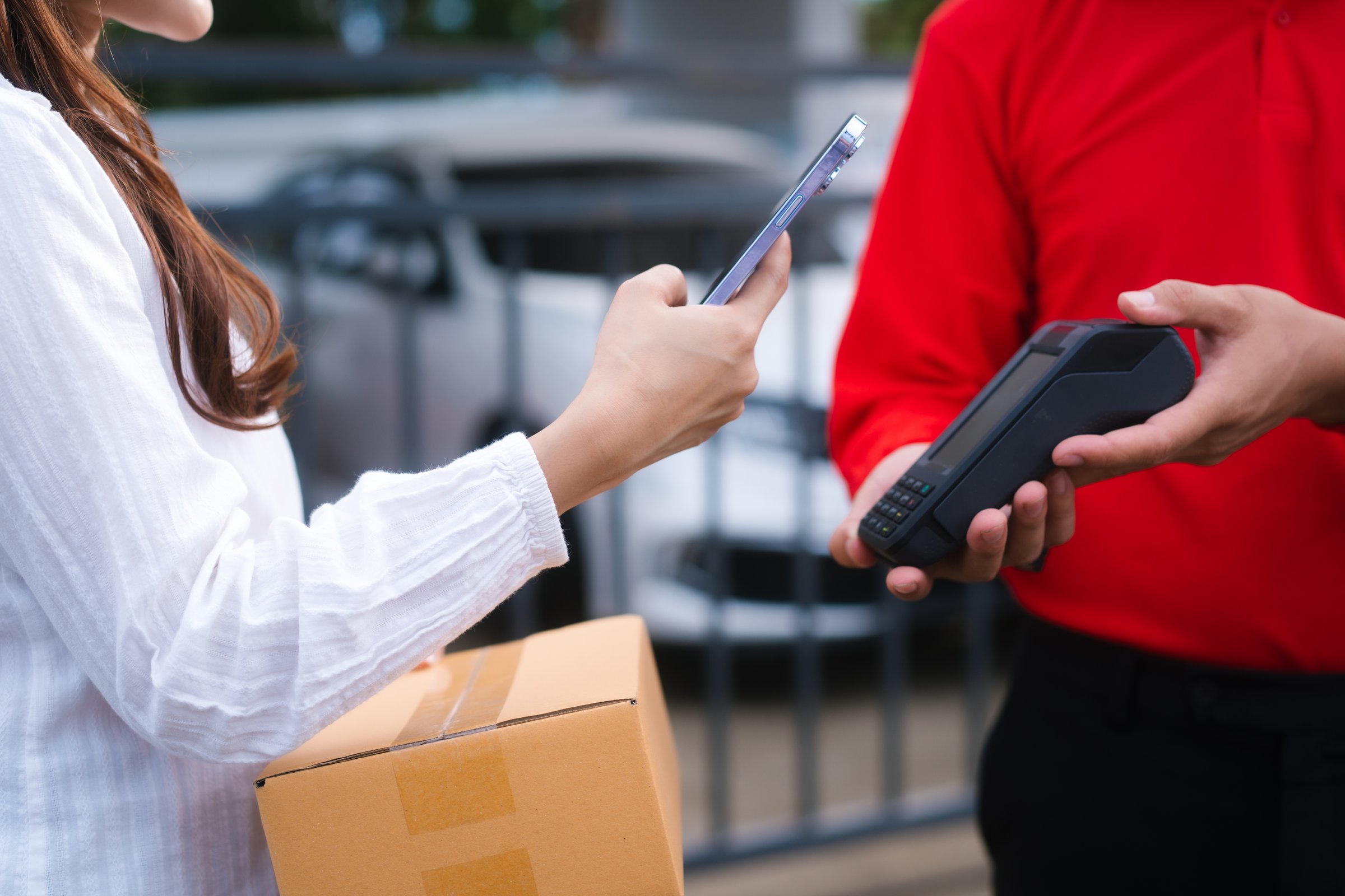 A delivery man, who appears to be Asian, in a red uniform hands a mobile payment terminal to a smiling woman, who seems to be middle-aged and holding a package, outside a modern home.