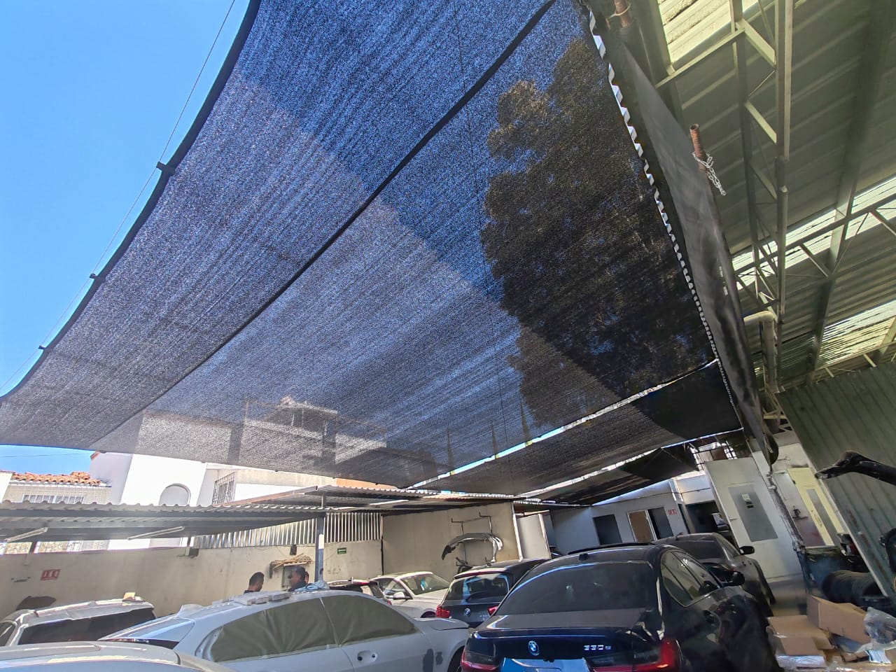 Shade cloths covering cars in a parking area, with sunlight filtering through and buildings visible in the background.