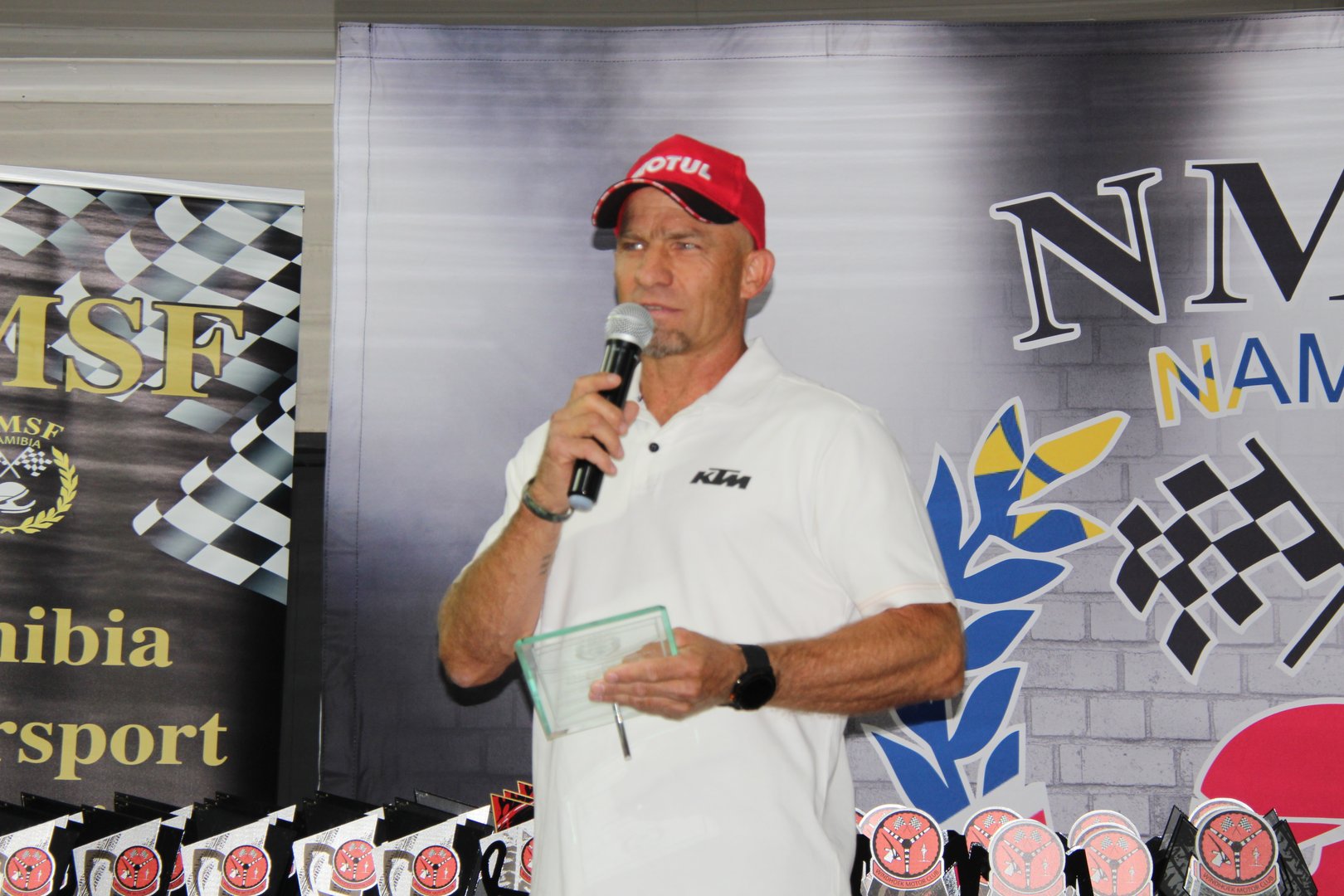 Man in white shirt and red cap speaks into a microphone at an event with banners and trophies in the background.