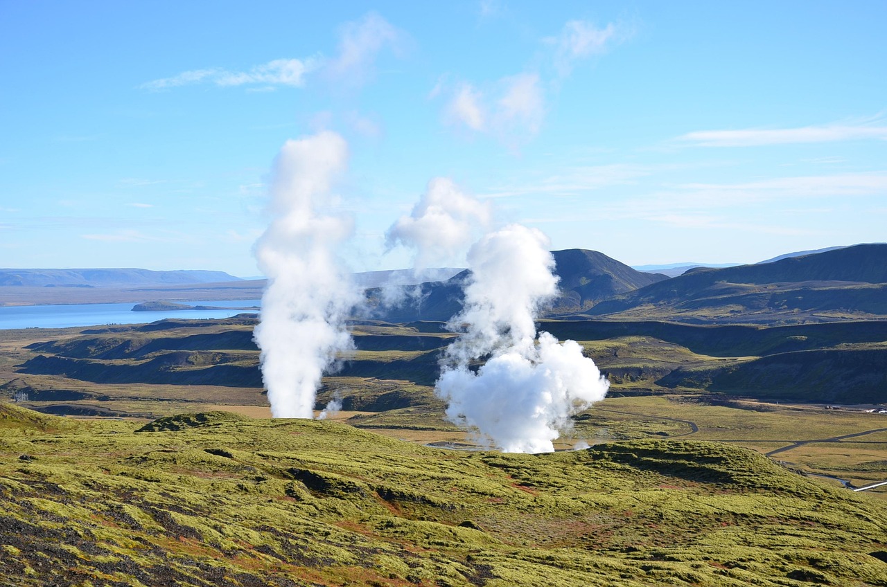 Geothermal steam vents in a mossy landscape with mountains and a lake in the background under a clear blue sky.