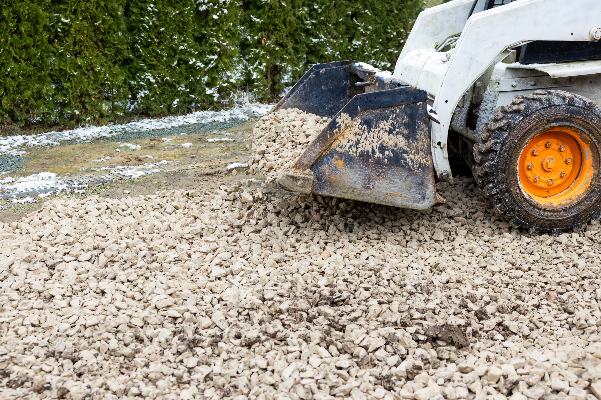 A small construction vehicle with orange wheels spreads light gravel on the ground. Evergreen hedges and patches of snow suggest a cold weather setting.