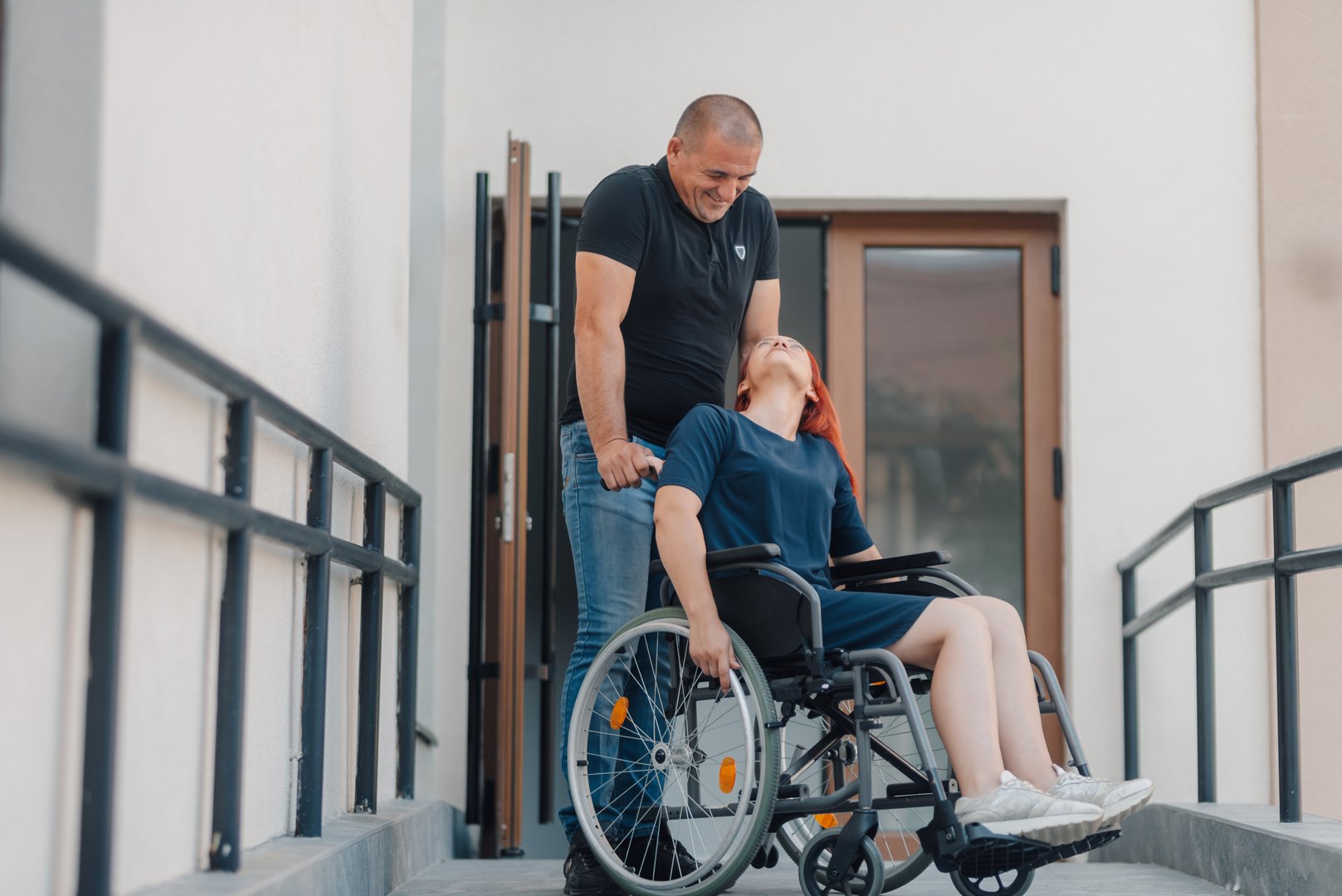 Man supports a smiling woman using a wheelchair as they navigate an accessible ramp, highlighting inclusivity and support