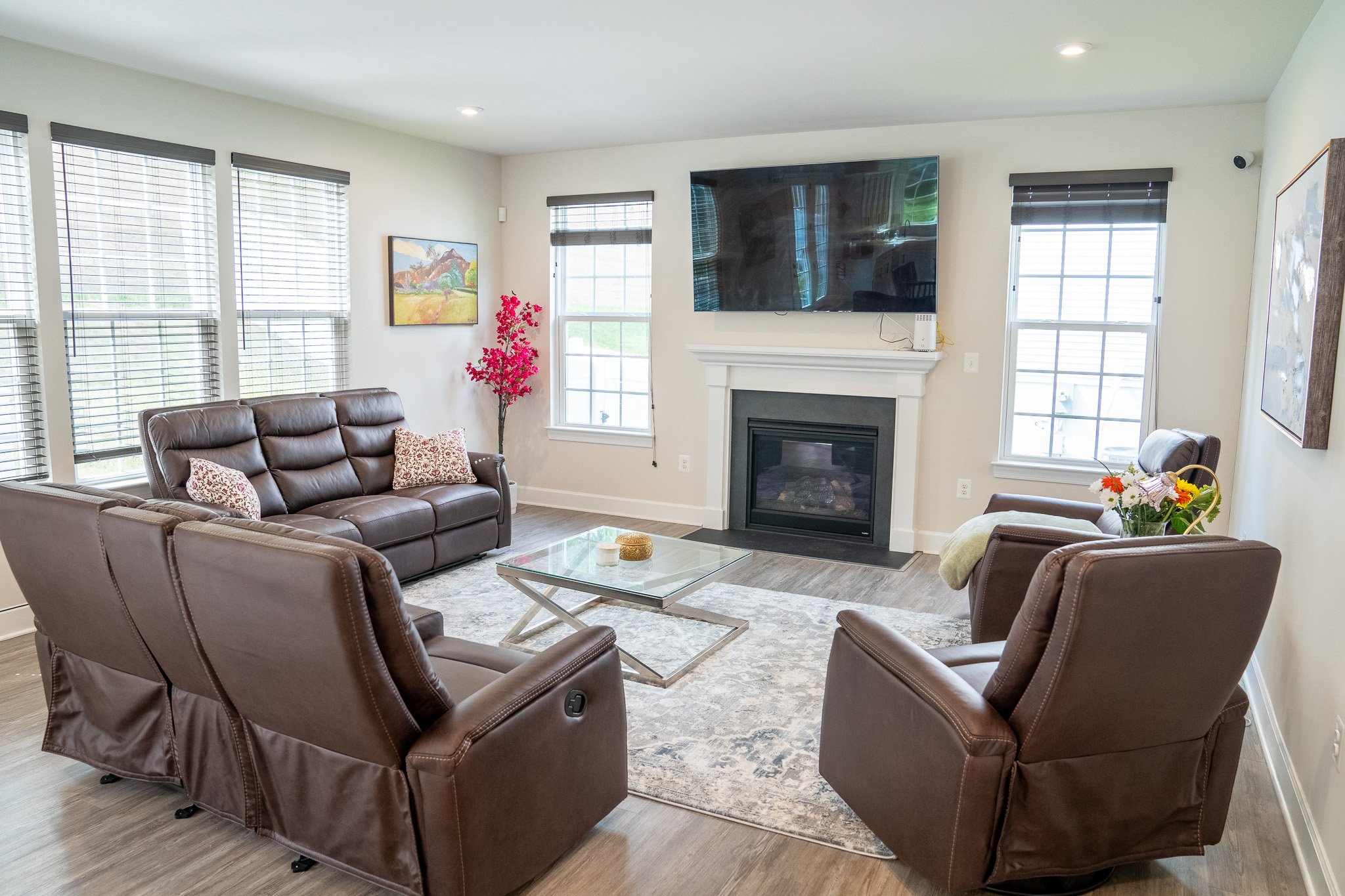 Modern living room with brown leather sofas, glass coffee table, fireplace, wall-mounted TV, and large windows.
