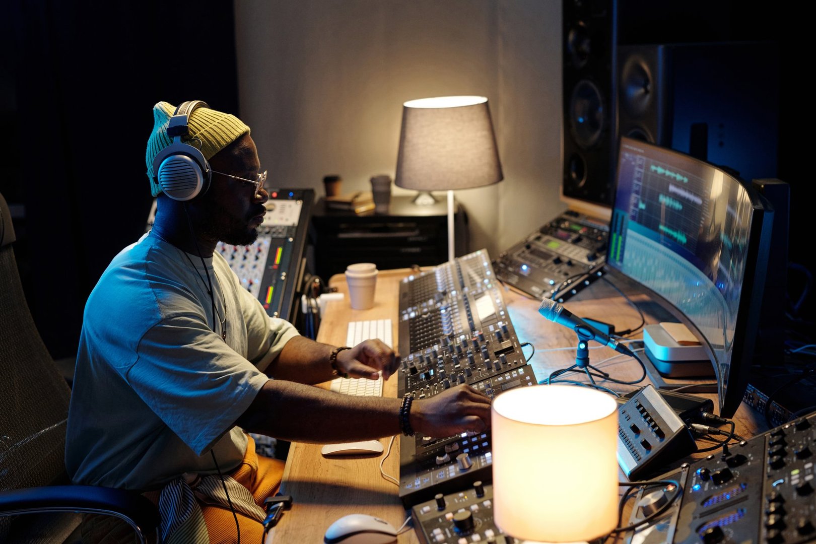 Man sitting at mixing console in studio, adjusting audio levels with concentration. Surrounded by professional audio equipment and computer screens, headphones on head, focused expression