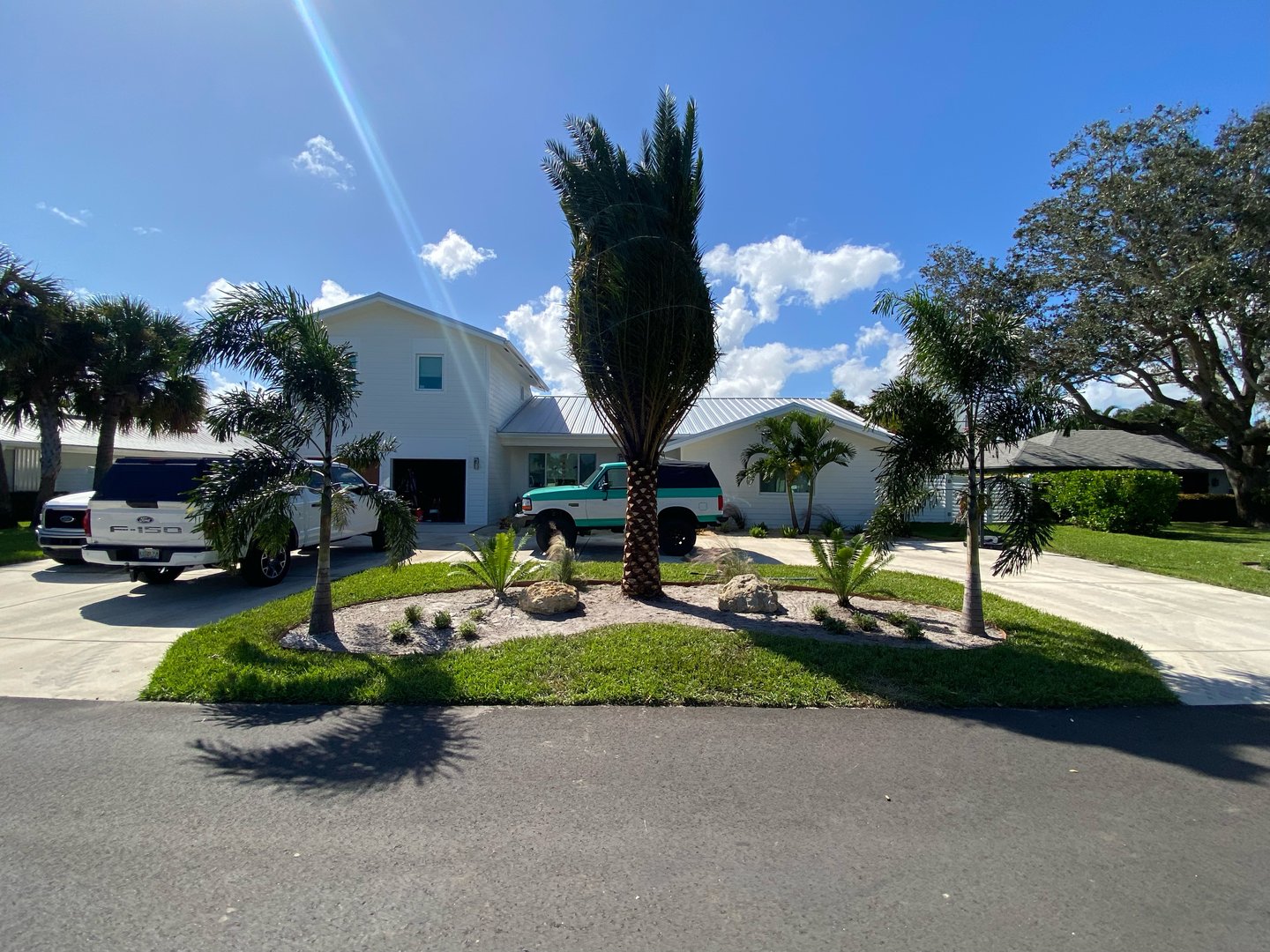House with white facade, two parked SUVs in driveway, palm trees and green lawn, under a bright blue sky.