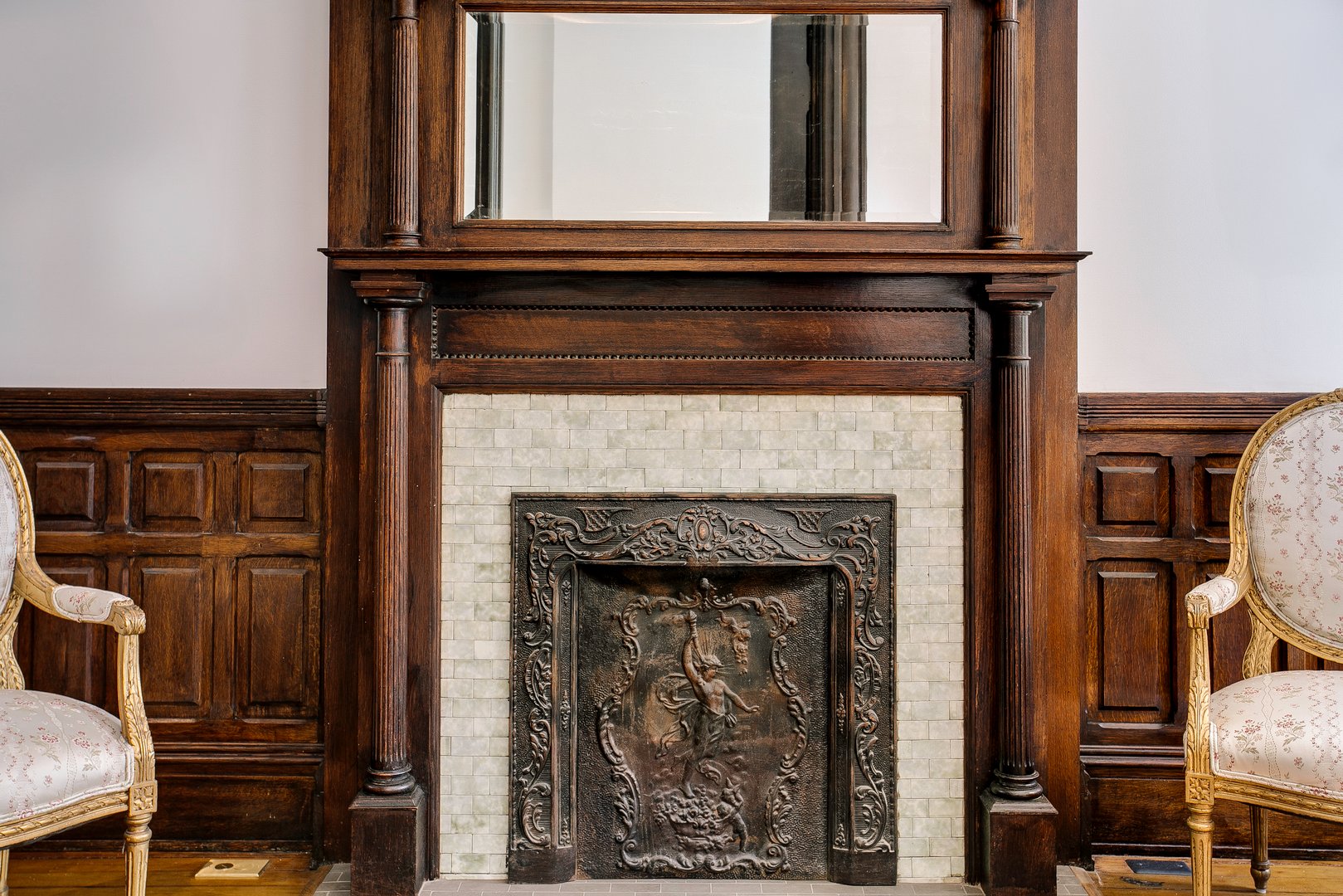 This image features an elegant vintage fireplace adorned with intricate metalwork, flanked by ornate wooden panels and classic upholstered chairs in an opulent interior setting.