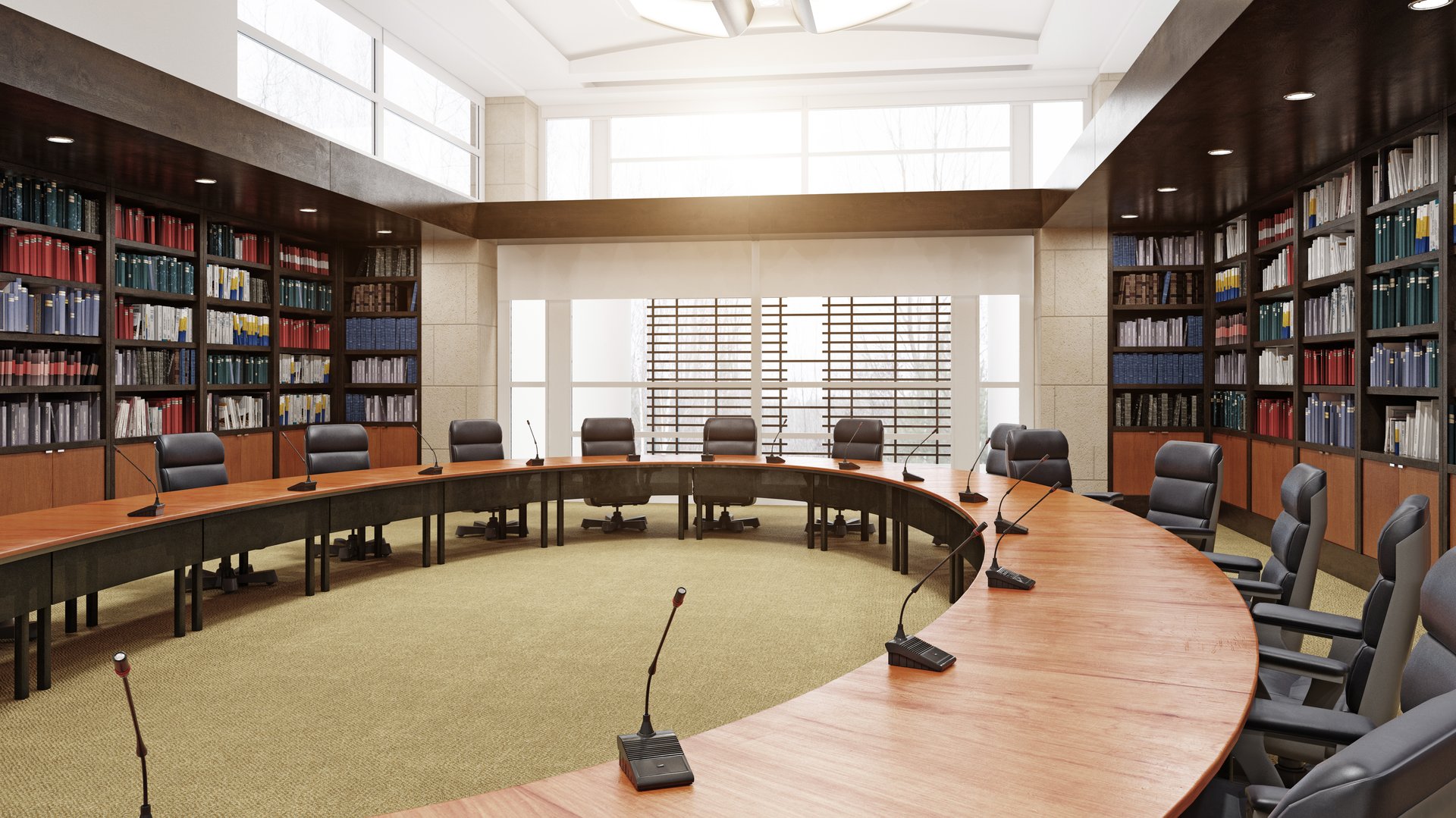 A 3D rendering of a modern, empty conference room with a large curved wooden table, black office chairs, and microphones. The room features floor-to-ceiling bookshelvesâ¦