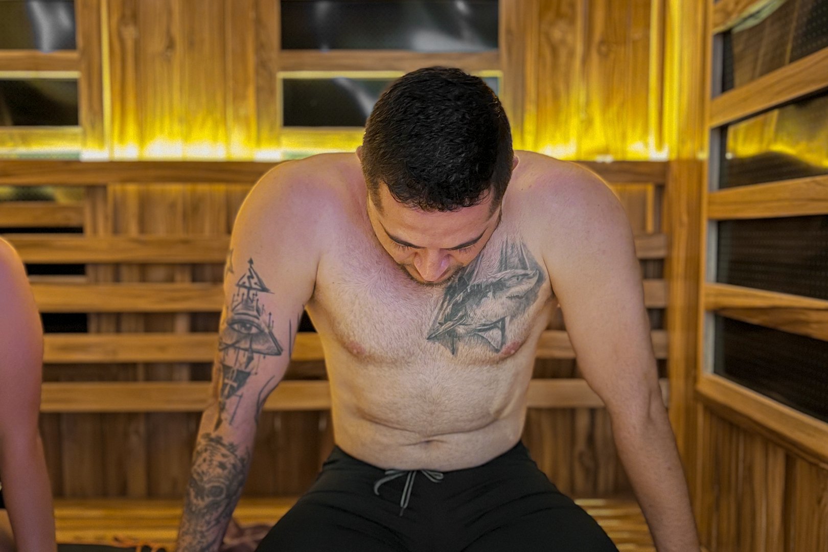 A man with tattoos on his chest and arm sits in a wooden sauna, looking down, with warm lighting in the background.