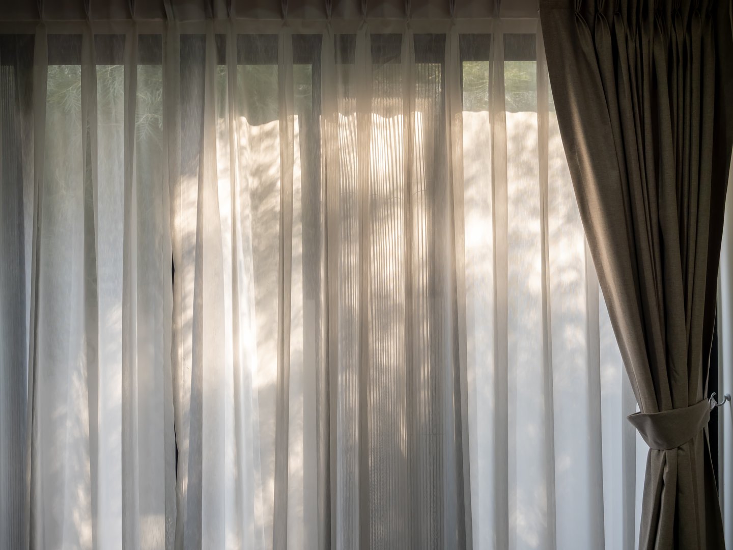 White transparent curtain at the window in the dark room with warm sunlight and tree leaves shade. Large horizontal window with closed white fabric curtain. Relaxation background concept.