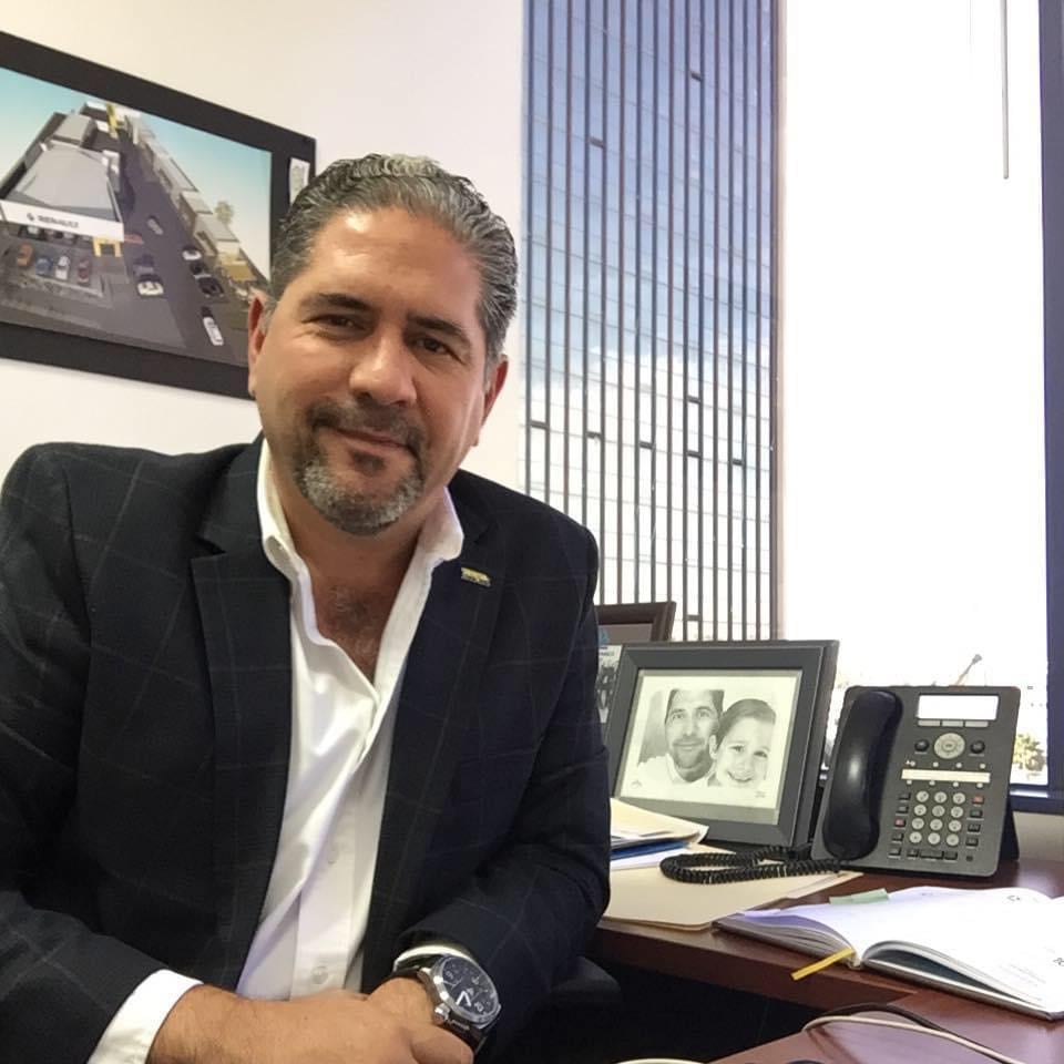 Man in a suit sitting at a desk with a telephone and framed photo. Large window view to city buildings behind him.