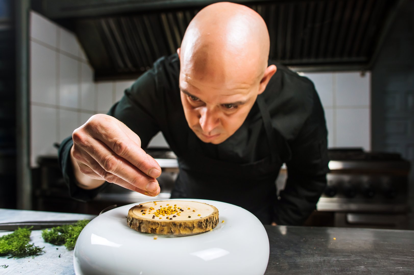 Young male bold chef dressed in black is flavouring a molecular dish. Dark background. Molecular cuisine