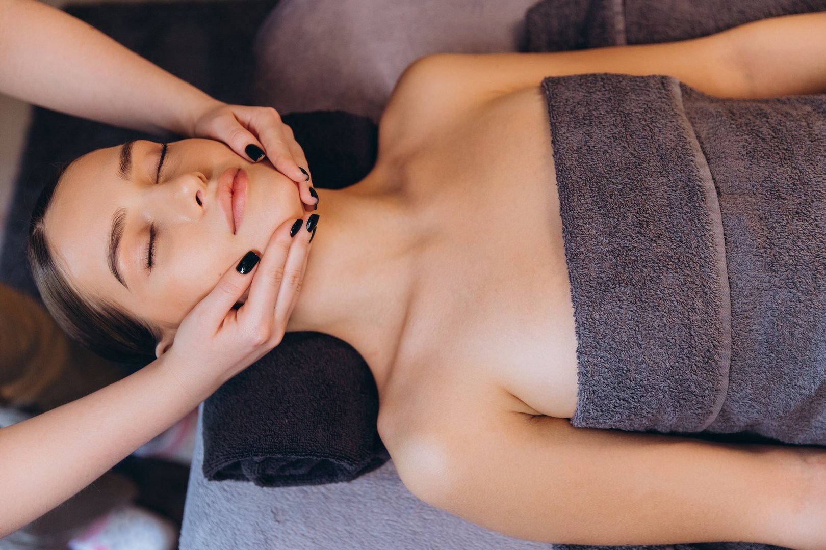 Massage therapist giving a facial massage to a young woman lying on a massage table with eyes closed, enjoying a moment of relaxation at the spa