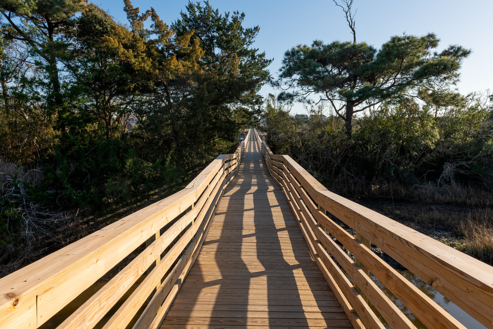 A long wooden footbridge stretches through coastal trees and marshland in Oak Island, North Carolina, under a clear blue sky.