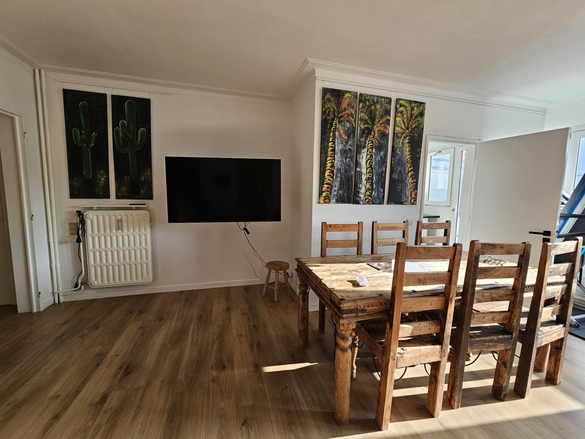 Dining area with wooden table and chairs, wall-mounted TV, and cactus paintings on a light-colored wall.