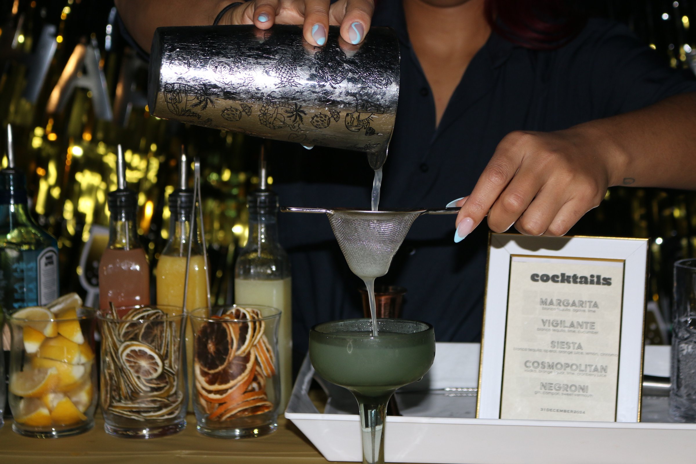 Bartender pouring cocktail through a strainer into a glass, surrounded by ingredients and a menu on a bar setup.