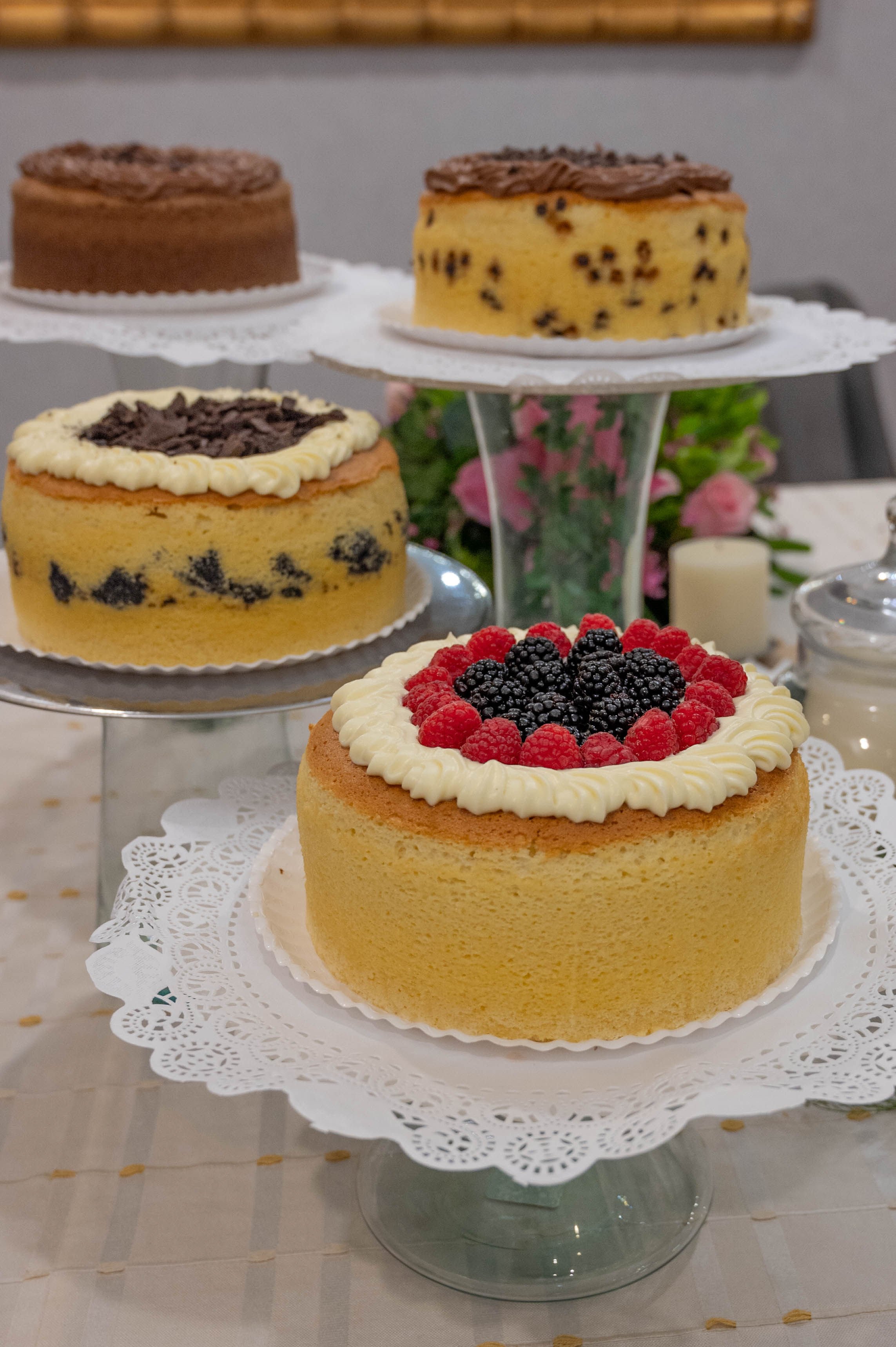 Four cakes on stands, each with different toppings like berries, chocolate chips, and cream, displayed on a table.