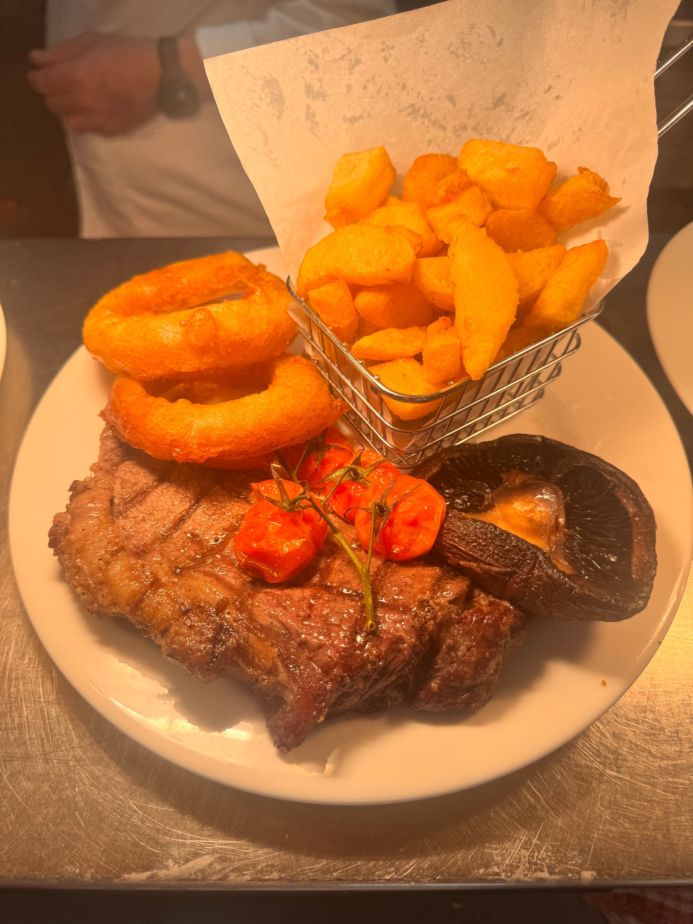 Grilled steak with crispy fries in a basket, onion rings, grilled tomatoes, and a mushroom on a white plate.