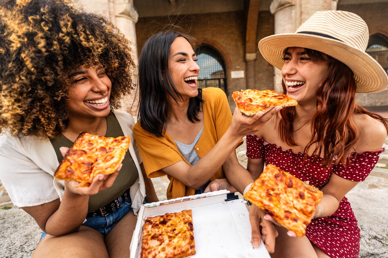 Three young female friends eating pizza sitting outside - Multiracial women enjoying street food in the city - Italian food culture and summer vacations concept
