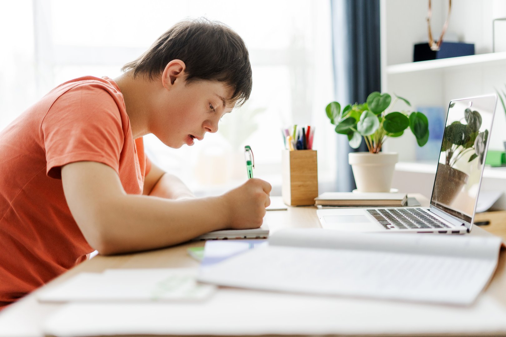 Focused teen boy with down syndrome writing in his notebook while studying at home, doing homework. Online education concept