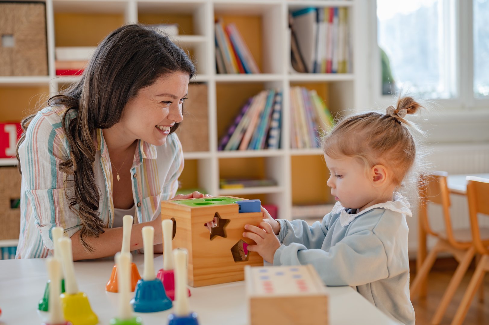 A young woman and a little girl play with educational toys at a preschool or daycare, engaging in learning activities with a shape sorter in a bright, cheerful classroom.