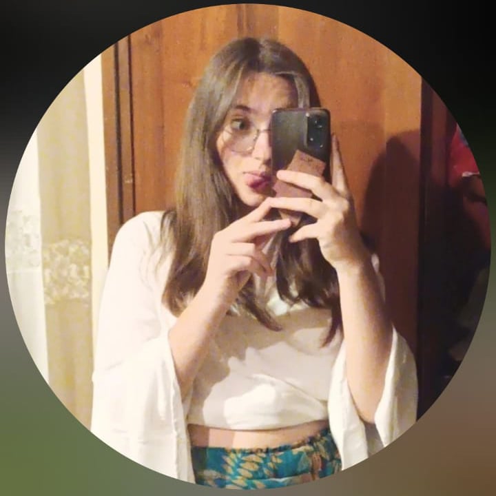 Young woman with glasses takes a mirror selfie, wearing a white long-sleeve top and floral pants.