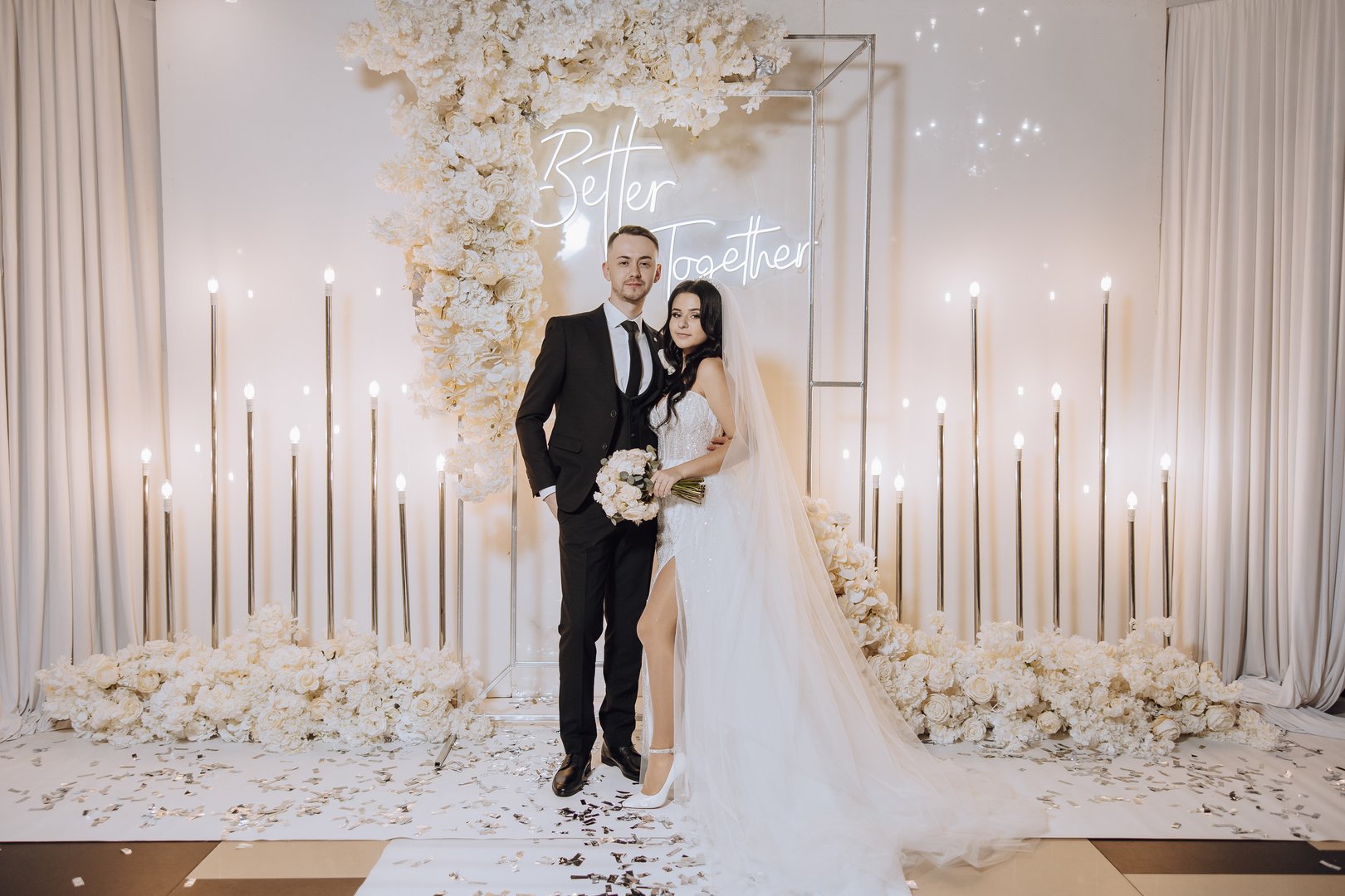 A bride and groom stand in front of a white backdrop with the words Better Together written on it