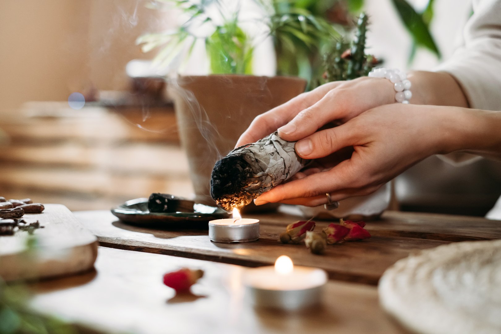Woman hands burning white sage, palo santo before ritual on the table with candles and green plants. Smoke of smudging treats pain and stress, clear negative energy