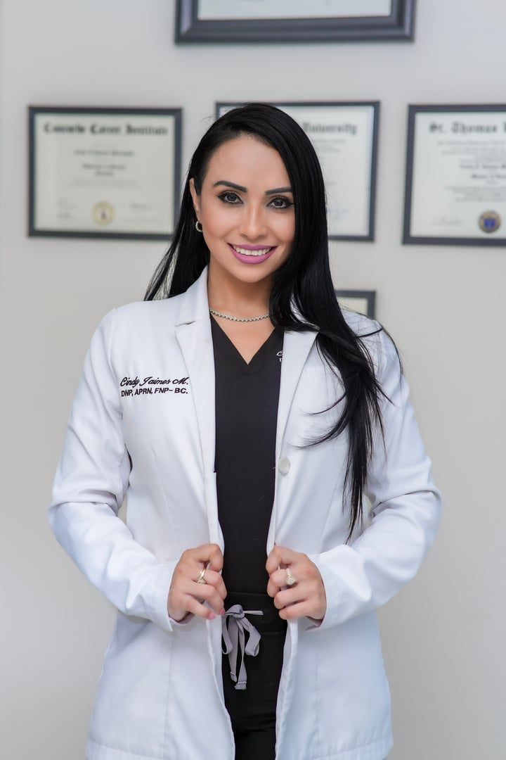 Smiling woman in a white lab coat standing in front of framed certificates on a wall.