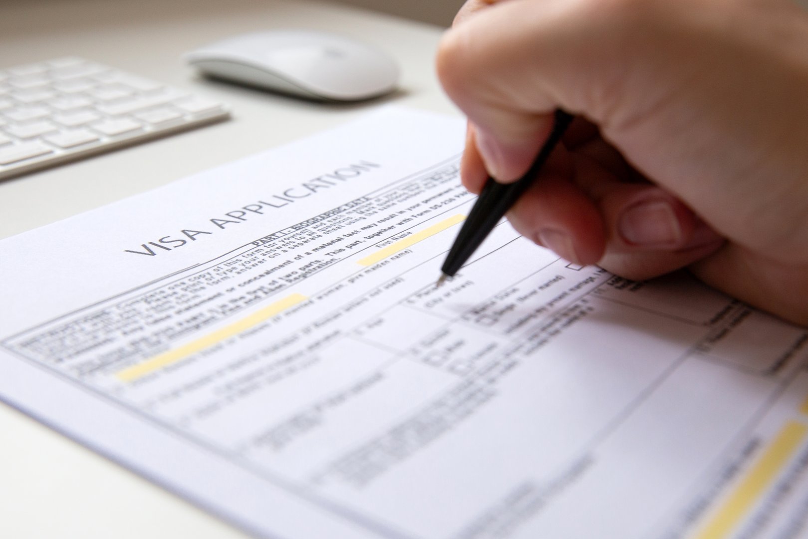 Close up of a female hand filling out a visa application document, white table with computer keyboard and a mouse in background