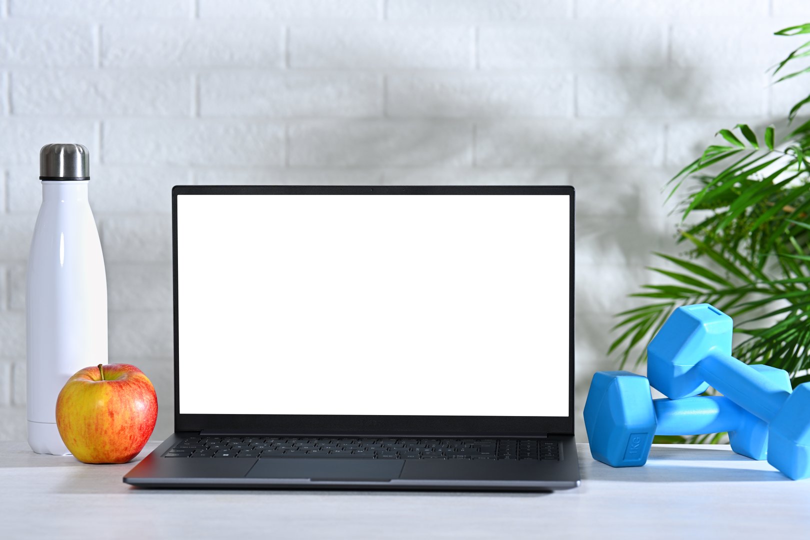 Laptop with white screen on table with dumbbells, bottle and apple. Online workout and healthy lifestyle concept
