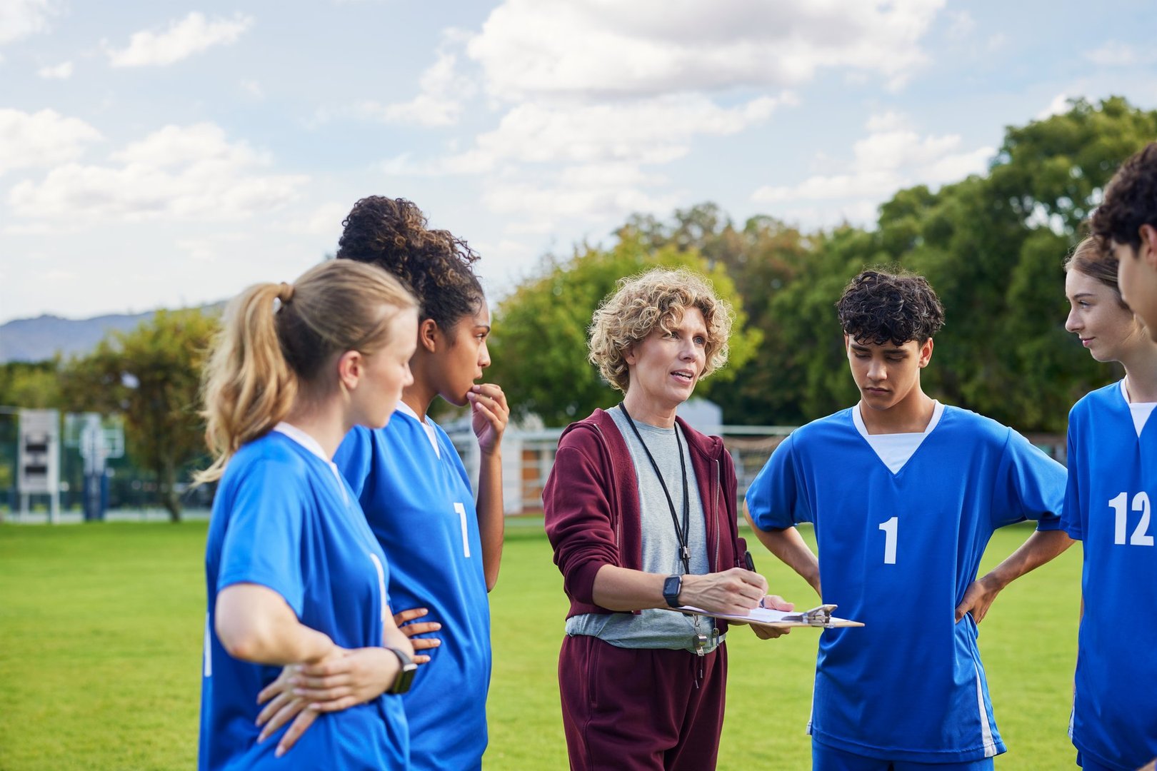 Focused female mature coach giving instructions to multiethnic players during a sports practice session. Coach sharing strategy and giving motivation to guys and girls players during match. Physical education teacher talking to sports teenagers or athlete group for workout at school lesson.