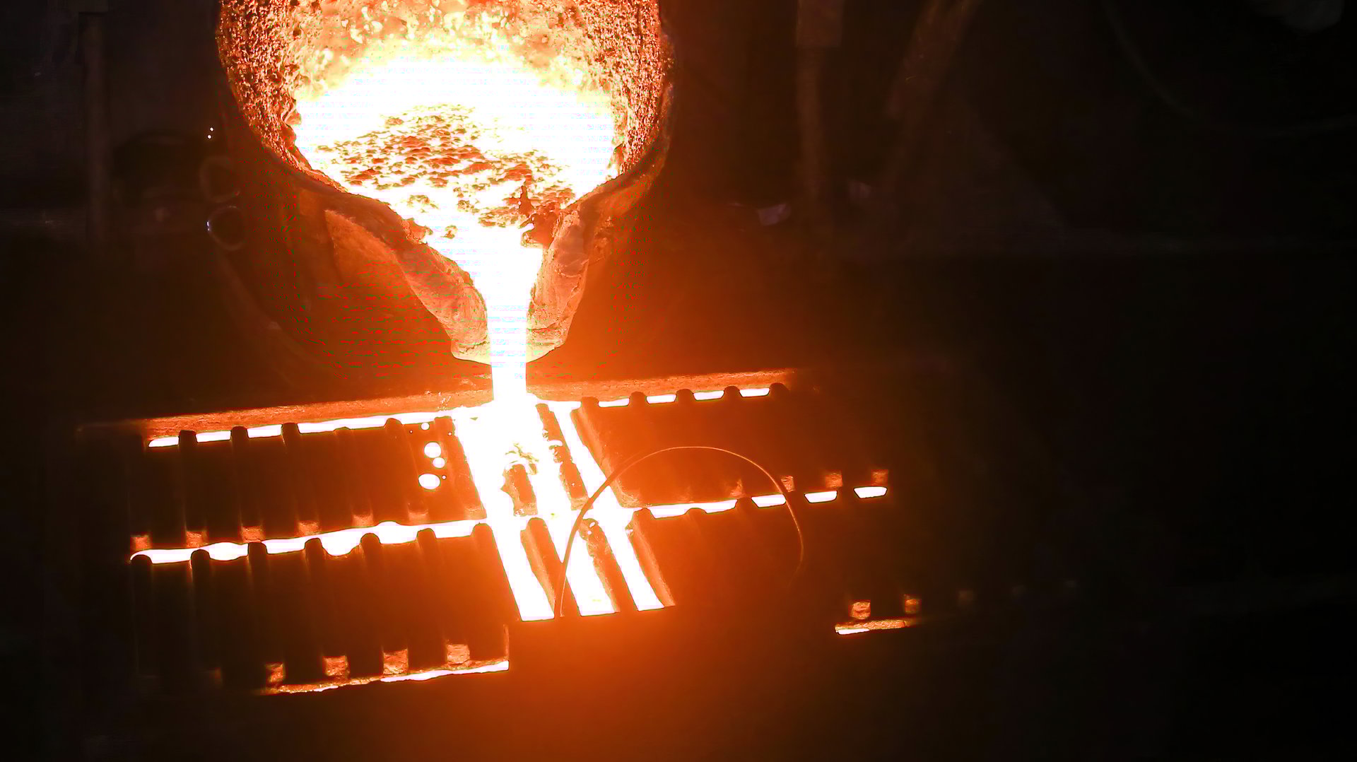 Molten metal poured from ladle into mold to make casting. Metal glows brightly, flows into mold, sparks fly as metal hits mold, on dark background, showcasing metal casting process.