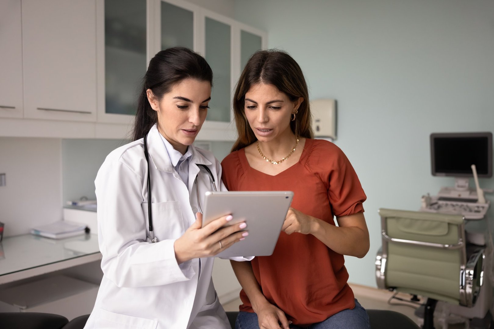 Focused young Latin doctor and female patient watching ultrasound scan shot on tablet, pointing at screen, talking, discussing examination result, medical report, electronic prescription