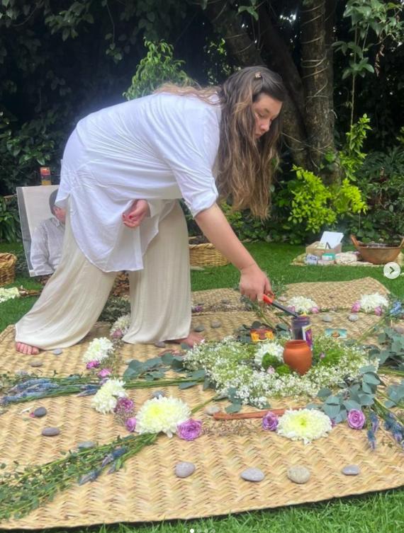 A woman in white clothing arranging flowers on a mat in a garden setting, surrounded by greenery and outdoor decor.