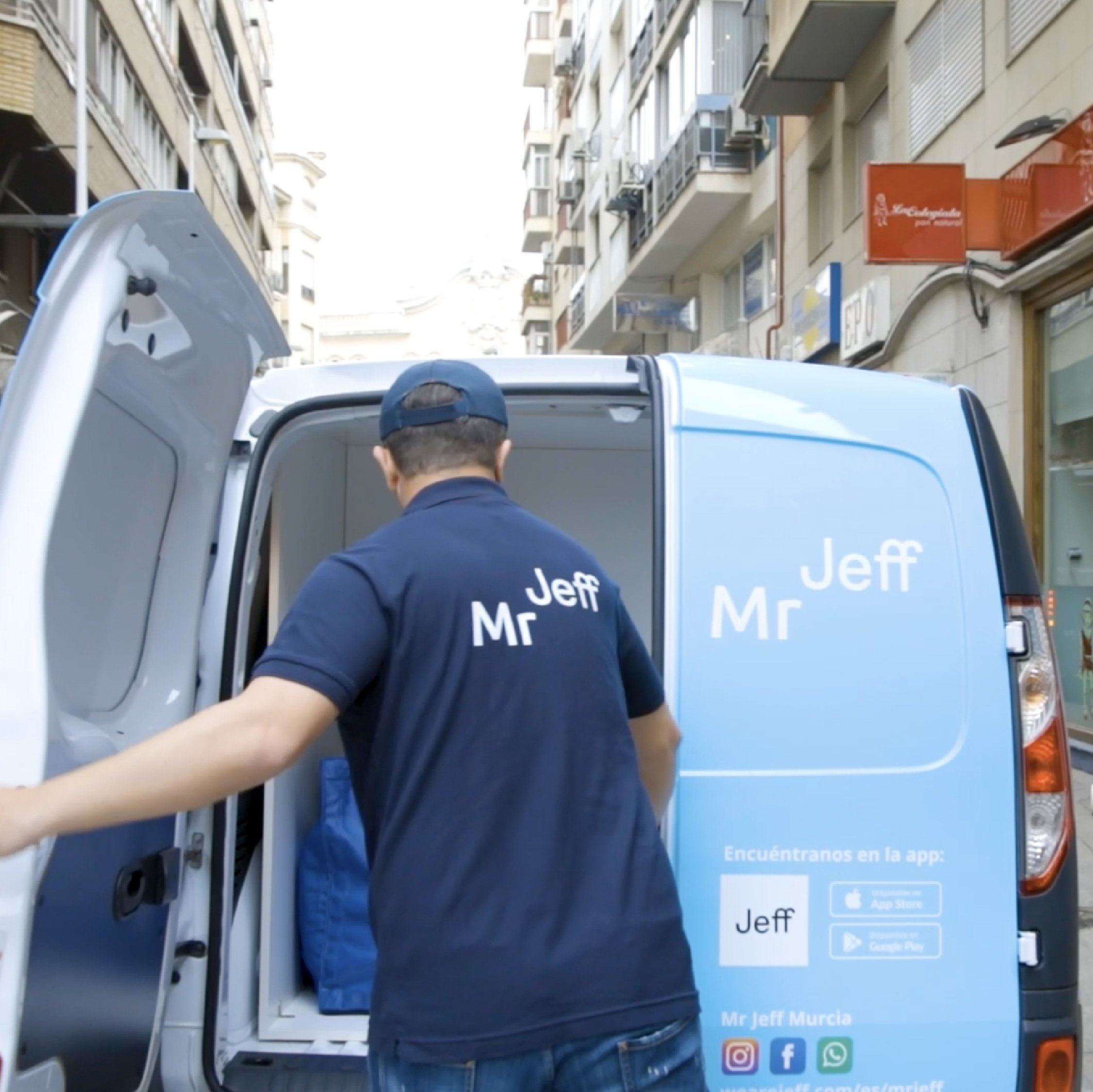 A delivery person wearing a Mr Jeff shirt unloads a blue van on a city street, next to buildings and signs.