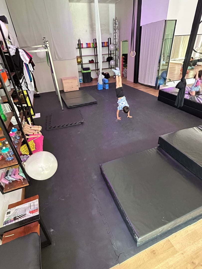 Child doing a handstand in a gym with padded flooring, surrounded by exercise equipment and mirrors on the walls.