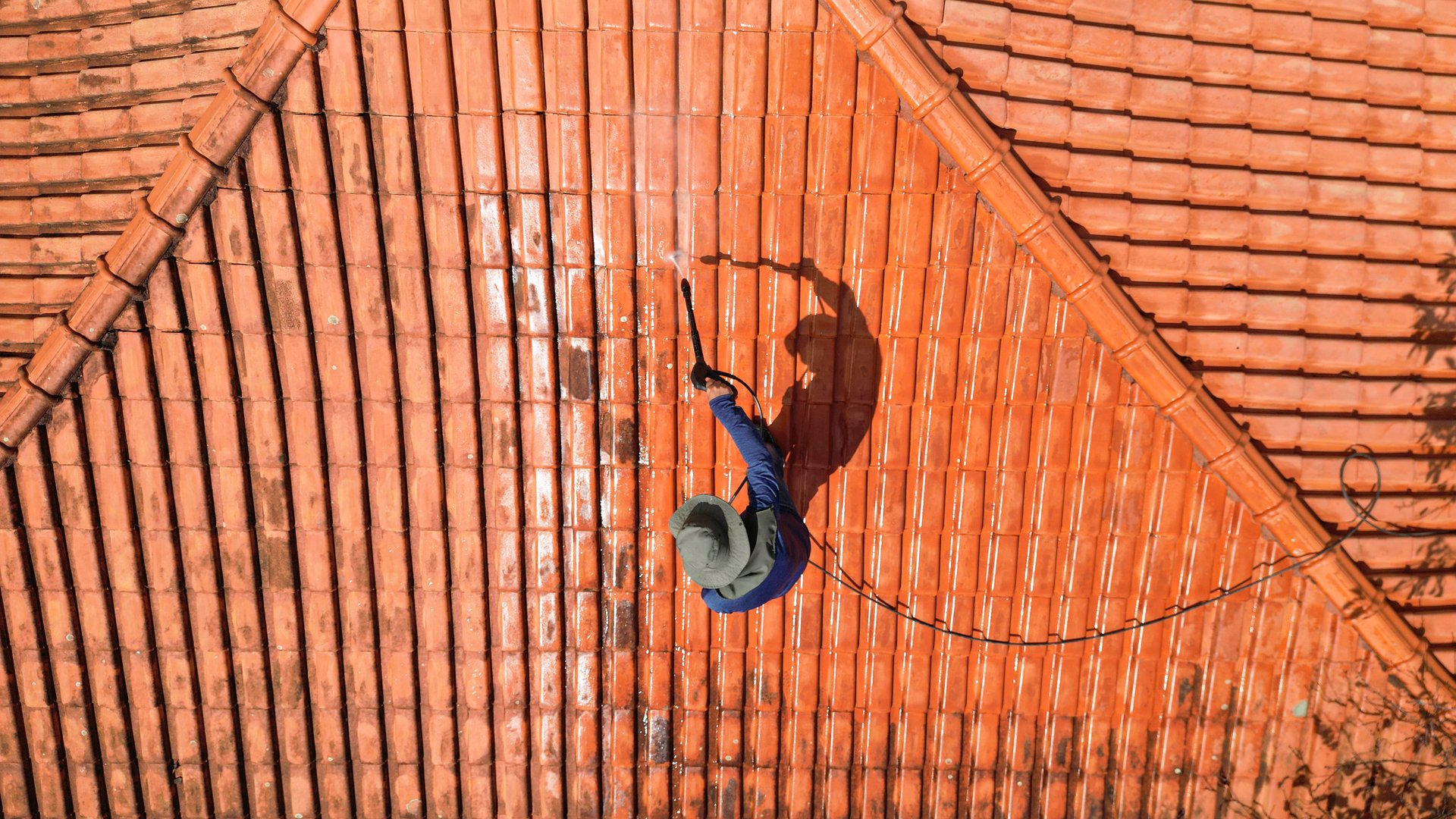 A person outdoors cleaning the red clay roof using a high pressure washer. Professional washing house tiles with water hose.
