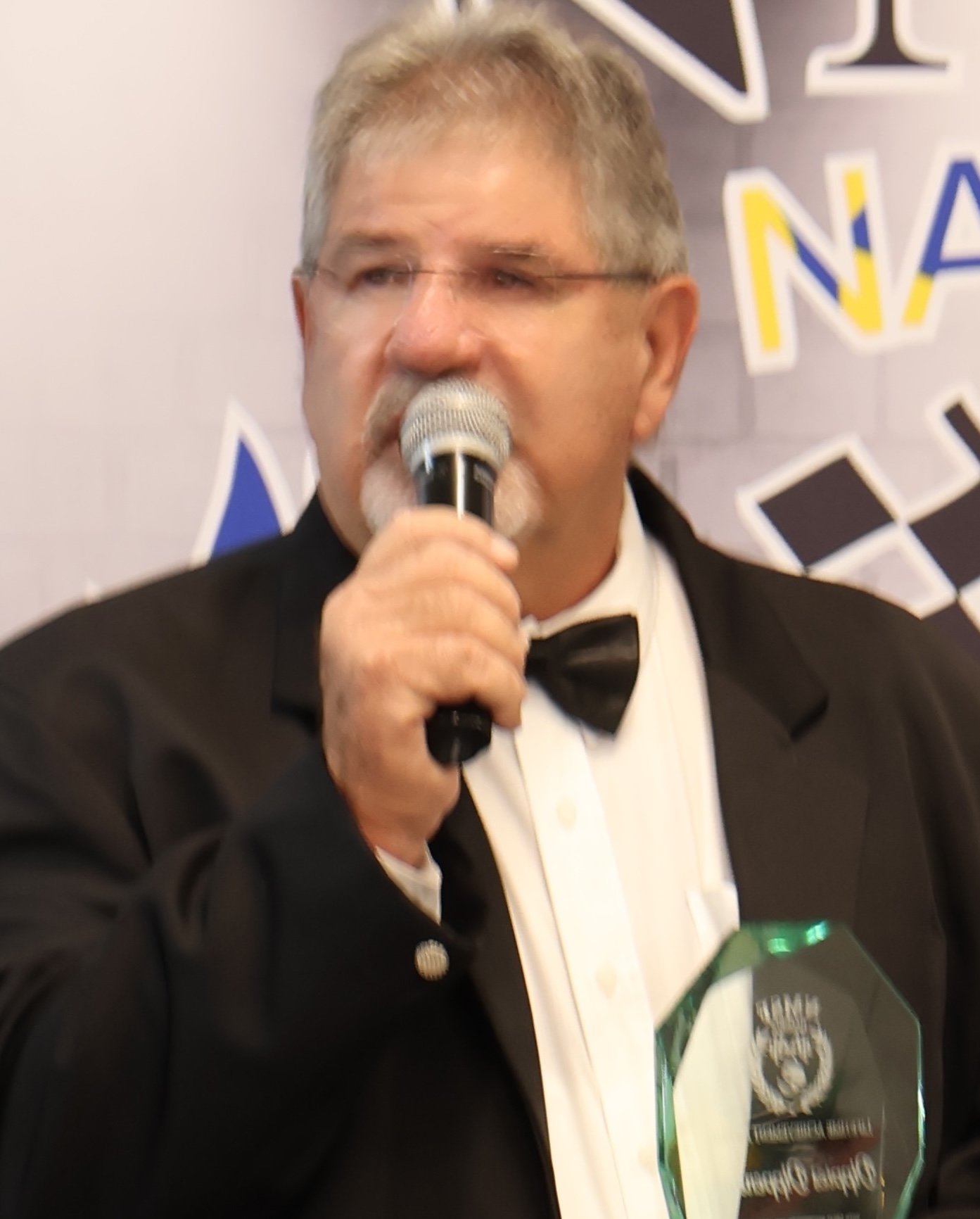 Man in a tuxedo holding a microphone and an award, speaking at an event with a blurred banner in the background.