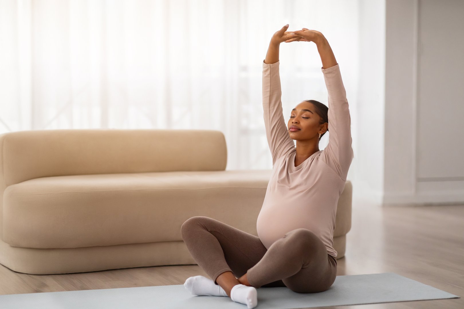 Peaceful young black pregnant woman have yoga at home, sitting on fitness mat with closed eyes at sunny living room, raising hands up. Workout during pregnancy, copy space