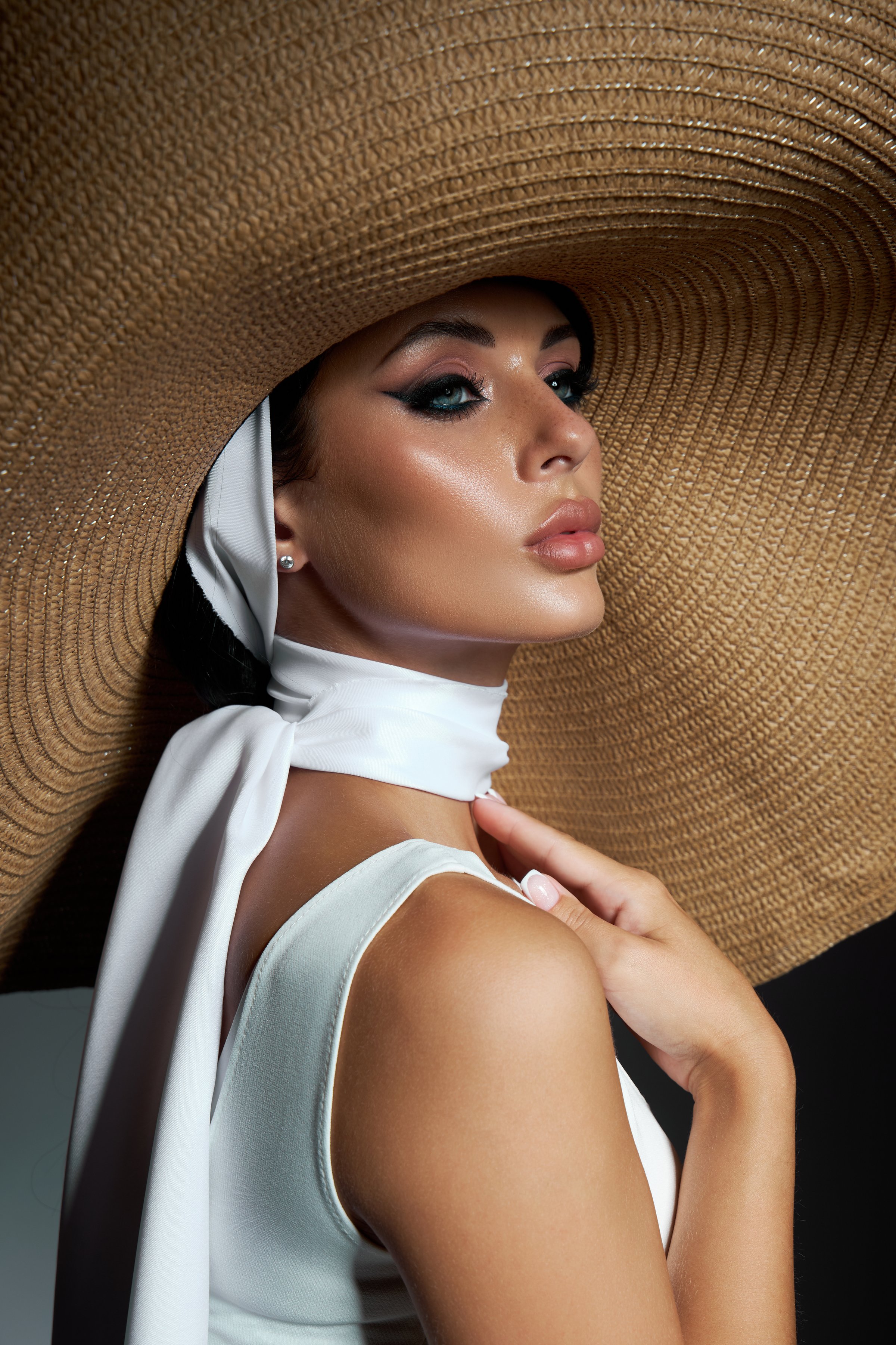 woman poses confidently, showcasing her striking makeup and fashion. large straw hat and flowing scarf highlight her graceful look within a professional studio.